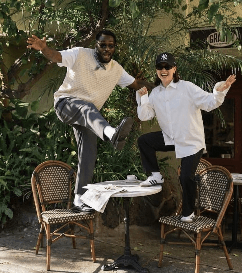 Two stylish people posing playfully at an outdoor café with tropical plants
